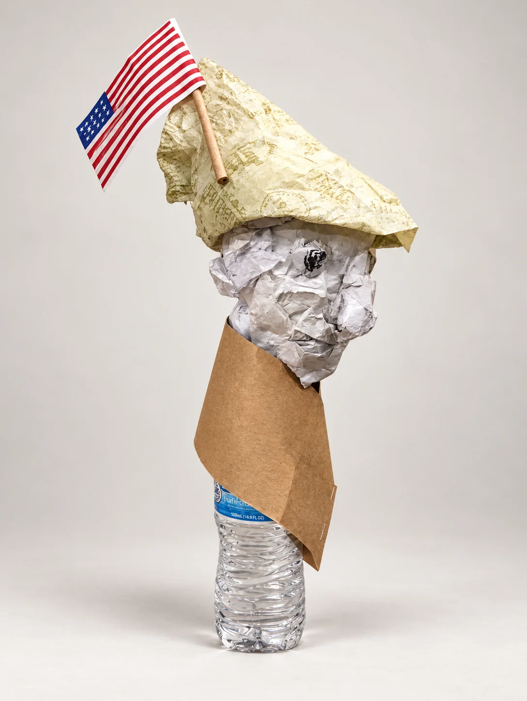 Untitled. Paper and found objects. A figure made from crumpled paper on a plastic water bottle, wrapped in brown paper and wearing a paper hat with a small American flag.