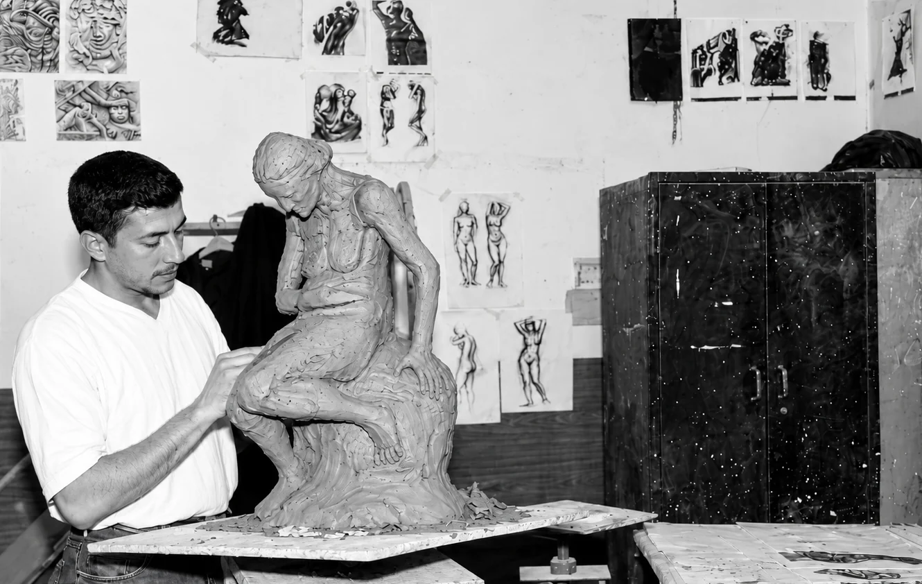 Mishaal Atheer modeling a seated figure in clay at the Baghdad Art Studio, 2002, with his drawings on the wall behind